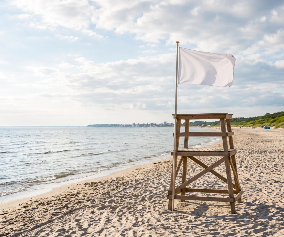Biała flaga na wieży ratowniczej przy pustej, piaszczystej plaży. O pogodzie w Międzyzdrojach przeczytasz na SE Szczecin.