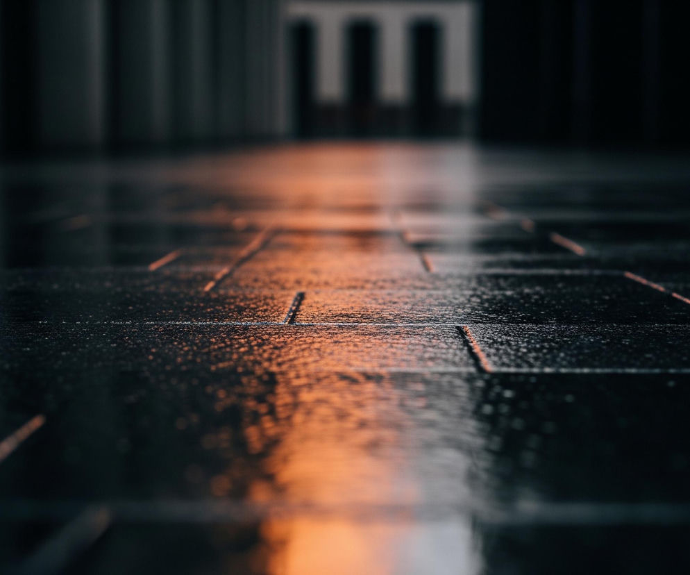 Wilgotna, ciemna posadzka, prawdopodobnie z płytek, rozciąga się w głąb obrazu, ukazując nieregularne, lśniące odbicia światła. Powierzchnia blisko obiektywu jest wyraźna i pokryta drobnymi kroplami wody, z wyraźnie widocznymi, równoległymi liniami fug. W oddali posadzka staje się rozmyta, a pomarańczowe światło tworzy szeroki pas rozjaśniający centrum kadru, zanikający ku górze obrazu. Po obu stronach widoczne są ciemne, pionowe kształty, przypominające kolumny lub ściany, a w tle majaczy niewyraźny zarys architektoniczny z prostokątnymi otworami.