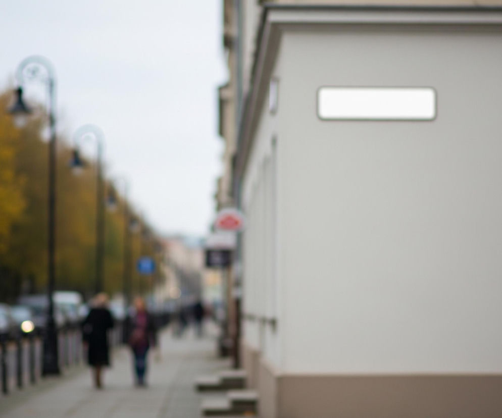 Na pierwszym planie po prawej stronie znajduje się kremowa ściana budynku z brązowym pasem u dołu i pustą, białą tabliczką z zielonkawą obwódką. Lewa część zdjęcia jest rozmyta, przedstawiając chodnik z czarnymi słupkami i lampami ulicznymi wzdłuż ulicy. W oddali widoczne są zaparkowane samochody oraz niewyraźne sylwetki ludzi idących chodnikiem, a za nimi drzewa o jesiennych liściach w odcieniach żółci i pomarańczu pod jasnym, zachmurzonym niebem.