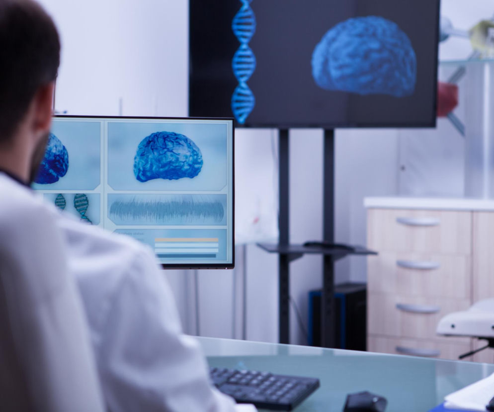Mężczyzna w fartuchu laboratoryjnym analizuje dane EEG i modele mózgu na monitorach, badając wpływ kofeiny na sen. Na ekranach widoczne są wykresy fal mózgowych oraz trójwymiarowe wizualizacje mózgu, co odzwierciedla badania naukowe, o których można przeczytać na portalu Poradnik Zdrowie.