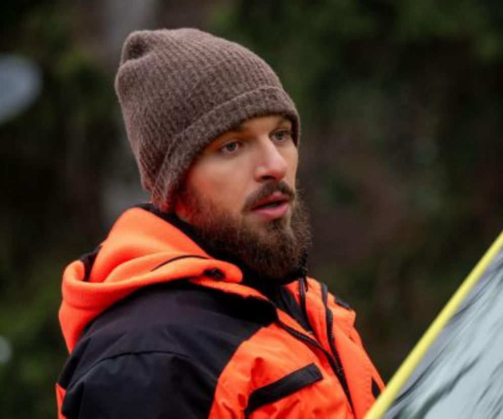 Mężczyzna w brązowej czapce i pomarańczowej kurtce ratownika medycznego, z brodą i zamyśloną miną. Zdjęcie ilustruje odcinek serialu Na sygnale, gdzie ratownicy walczą o życie pacjentów w Leśnej Górze. Więcej o serialu przeczytasz na Super Seriale.