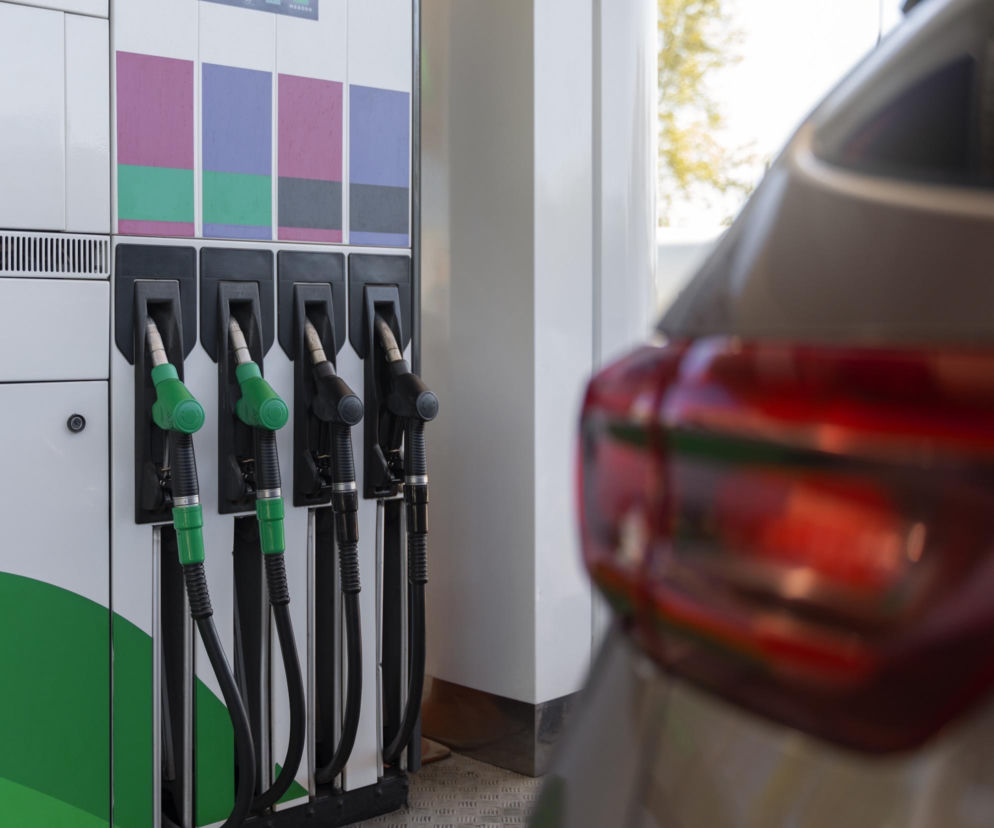 Dystrybutor paliwa z pistoletami, na pierwszym planie samochód z czerwoną lampą tylną. Widoczne zielone i czarne uchwyty pistoletów. Zgodnie z informacjami na Super Biznes, fiskus kontroluje stacje benzynowe za zawyżanie cen paliw, grożąc wysokimi karami.