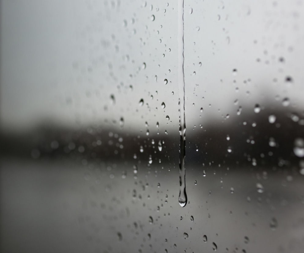 Szyba pokryta licznymi kroplami deszczu, z której spływa długa smuga wody, dominującej w środkowej części obrazu. Tło jest rozmyte i szare, sugerując pochmurną pogodę lub scenę widzianą przez szybę w deszczu. Po lewej stronie widoczne są ciemne, pionowe pasy, prawdopodobnie fragment ramy okna lub futryny, natomiast reszta tła jest nieostra i ma odcienie szarości oraz bieli.