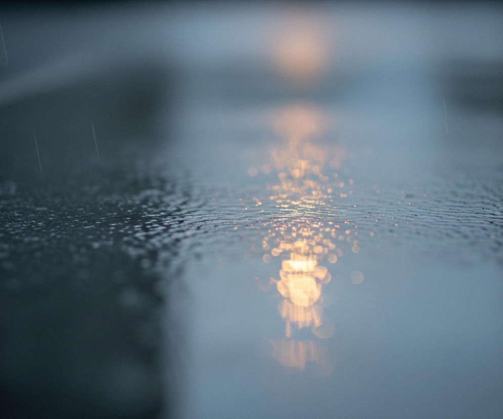 Ulica pokryta wodą odbija światła, tworząc rozmyte, pomarańczowo-żółte smugi wzdłuż środkowej części kadru. Krople deszczu są widoczne na mokrej powierzchni asfaltu, szczególnie w lewej, górnej części obrazu. Całość utrzymana jest w chłodnych odcieniach szarości i błękitu, z wyraźnym kontrastem rozświetlonych refleksów.