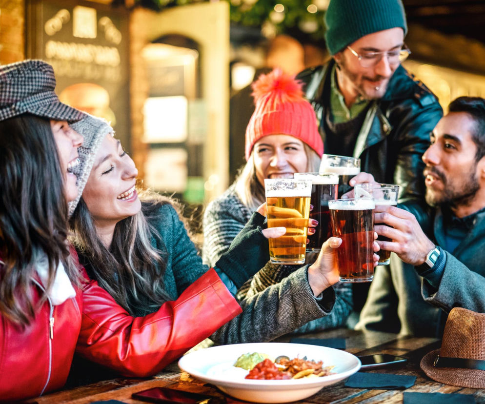 Grupa przyjaciół w czapkach, śmiejąc się, wznosi toasty piwem, siedząc przy drewnianym stole w restauracji. Talerz z jedzeniem i kapelusz leżą na stole. Dowiedz się więcej o codziennym piciu piwa i jego wpływie na organizm na portalu Poradnik Zdrowie.