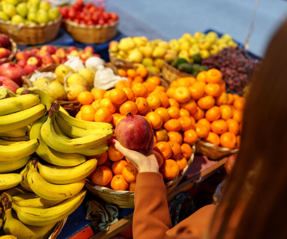 Ręka osoby trzymającej owoc granatu na tle straganu z bananami i pomarańczami. O diecie na nadciśnienie przeczytasz na PZ Fit.