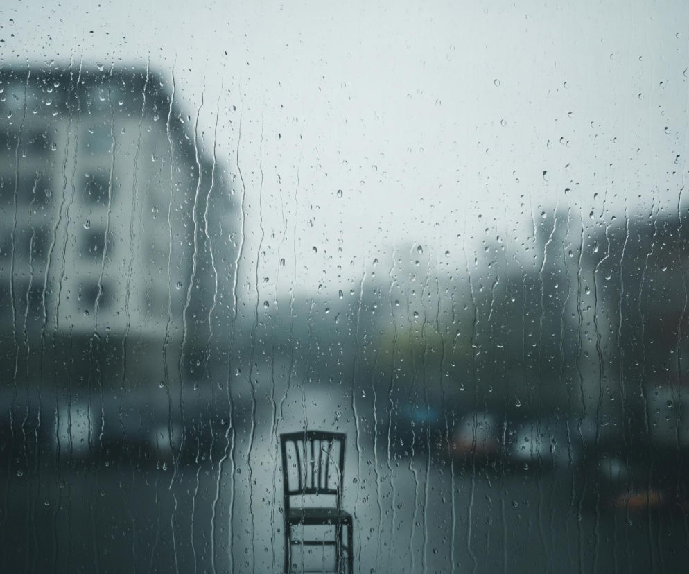 Widok zza mokrej szyby, na której znajdują się liczne krople deszczu i smugi wody, zamazujące tło. Na pierwszym planie, w centralnej dolnej części, widoczne jest czarne krzesło, ustawione przodem do obserwatora. Tło to rozmyty pejzaż miejski, z widocznymi zarysami budynków o jasnoszarych i ciemniejszych odcieniach, oraz niewyraźną ulicą poniżej. Cała scena utrzymana jest w chłodnych, szaro-niebieskich barwach, sugerujących pochmurny, deszczowy dzień.