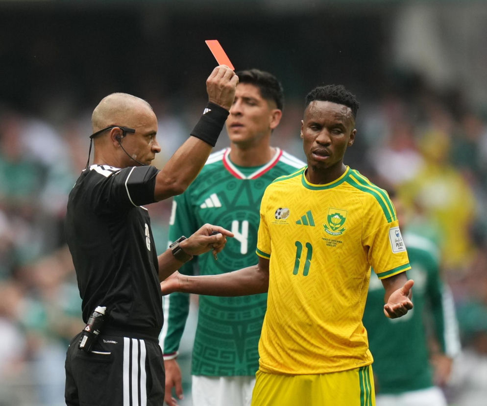 Sędzia pokazuje czerwoną kartkę Thembi Zwane, piłkarzowi w żółtej koszulce z numerem 11, po faulu na Roberto Alvarado. Zwane ma zrezygnowaną minę, a w tle widać zawodnika Meksyku. O surowej karze dla zawodnika przeczytasz na naszym portalu.