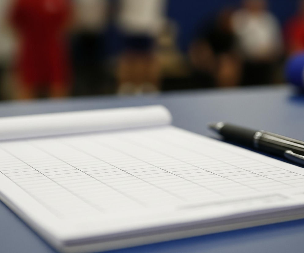 Na niebieskim stole leży otwarty notes z białymi kartkami w kratkę, obok niego znajduje się czarny długopis z metalowymi akcentami. Notes jest skierowany do przodu, a długopis leży ukośnie, częściowo na notesie, częściowo na stole. Tło jest rozmyte, z widocznymi zarysami postaci w czerwonych i białych koszulkach, oraz fragmentami niebieskich i czarnych powierzchni. Po prawej stronie w tle widać również stos zwiniętych białych arkuszy papieru z niebieskim elementem.