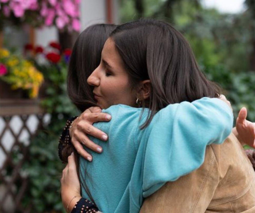 Agnes (Amanda Mincewicz) i Basia (Karina Woźniak) obejmują się na pożegnanie, zanim Agnes ucieknie z Grabiny przed prześladowcą, o czym przeczytasz więcej na Super Seriale. W tle widać zieleń i kwiaty.