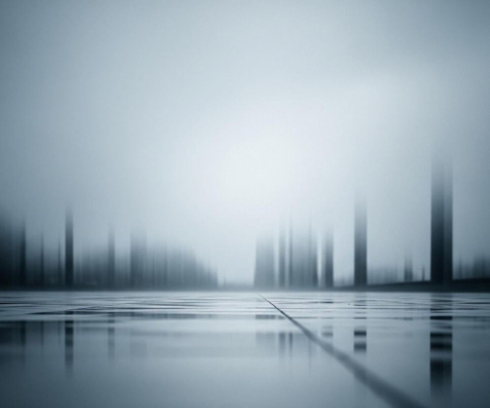 Widoczna jest perspektywa z poziomu gruntu, przedstawiająca podłogę wyłożoną jasnoszarymi kwadratowymi płytkami, z wyraźną linią fugi biegnącą przez środek. Powierzchnia jest mocno odbijająca światło, sprawiając wrażenie mokrej lub bardzo gładkiej, co tworzy lustrzane odbicia pionowych, rozmytych kształtów w tle. Te ciemniejsze, pionowe formy przypominające wieżowce lub słupy, rozciągają się w głąb obrazu, stopniowo zanikając w jasnej, zamglonej przestrzeni horyzontu. Całość utrzymana jest w chłodnych, monochromatycznych odcieniach szarości i błękitu, z dominującą bielą w centralnej części górnego planu, co potęguje wrażenie głębi i nieokreśloności.