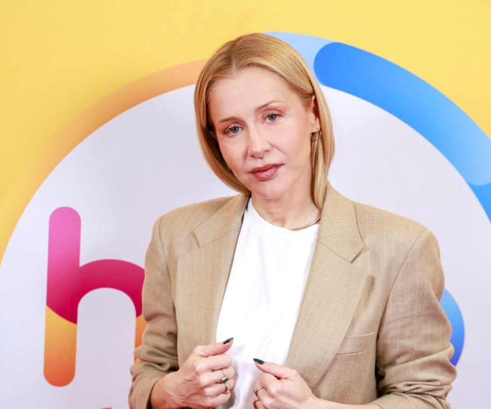 Aktorka Katarzyna Warnke w beżowej marynarce na tle kolorowego logotypu. O jej metamorfozie przeczytasz na SE.