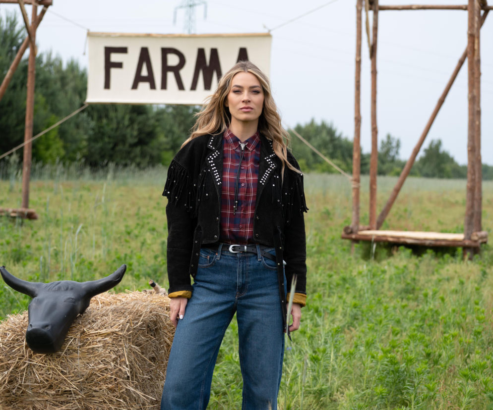 Kobieta w stylu country, w czarnej kurtce z frędzlami, koszuli w kratę i jeansach, pozuje na tle zielonego pola z napisem Farma i balotem słomy z figurką byka. Zbliża się finał Farmy z dodatkowym tytułem. Więcej o tym przeczytasz na Super Seriale.