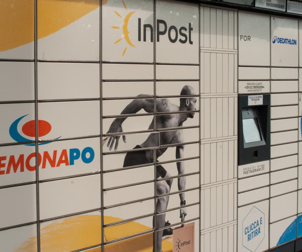 Paczkomat InPost we Włoszech z logotypami Cremonapo i Decathlon, reklamami biegacza oraz kobiety ćwiczącej, informuje o możliwości nadania paczki. O zasadach wysyłki InPost z Włoch do Polski przeczytasz na Super Biznes.