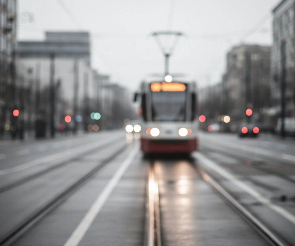 Na pierwszym planie, centralnie, widać tory tramwajowe biegnące przez całą długość szarej, mokrej jezdni. W oddali, na końcu torów, zbliża się niewyraźny tramwaj w kolorze białym z czerwonym pasem u dołu, z wyraźnie świecącymi, białymi reflektorami. Po obu stronach ulicy rozciągają się rozmyte, wysokie budynki miejskie, w większości w odcieniach szarości, z kilkoma rozproszonymi, czerwonymi i zielonymi światłami. Cała scena jest zamglona i utrzymana w chłodnej, szarej palecie barw, co tworzy wrażenie pochmurnego dnia lub zmierzchu.
