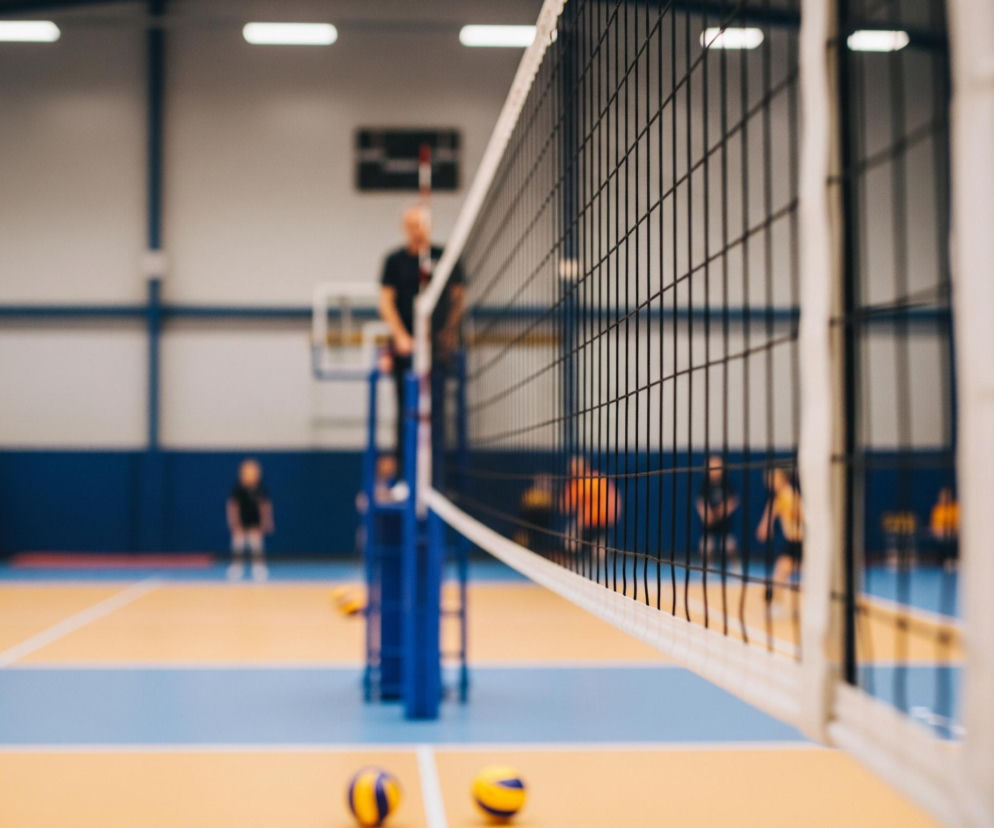 Na pierwszym planie widoczna jest siatka do siatkówki, rozciągnięta w poprzek kadru, z wyraźnie widocznymi czarnymi oczkami i białym pasem na górze. Pod siatką leżą dwie żółto-niebieskie piłki do siatkówki, częściowo niewyraźne. Dalej, w tle, znajduje się hala sportowa z żółto-niebieskim parkietem i białymi liniami, a także niewyraźne postacie ludzi i fragmenty wyposażenia sportowego, w tym niebieskie słupki podtrzymujące siatkę.