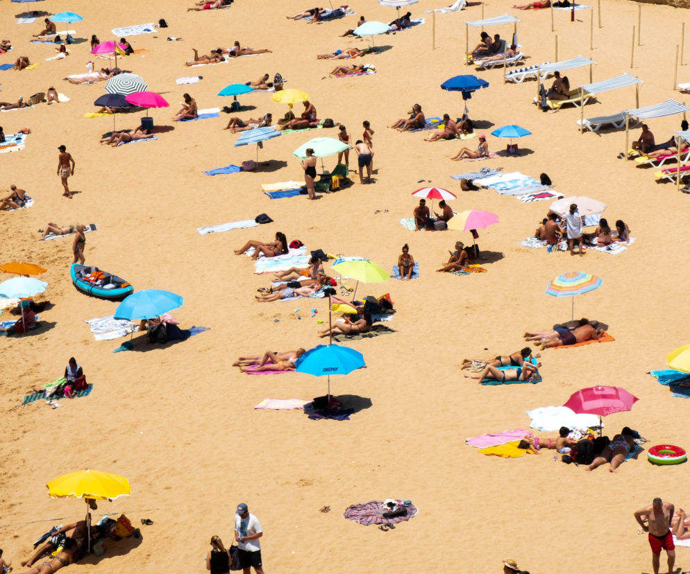 Plaża pełna ludzi odpoczywających na piasku pod kolorowymi parasolami, leżakami i zadaszeniami, idealne miejsce na upalny weekend, o którym przeczytasz więcej na naszym portalu. Słońce świeci mocno, a w tle widać piaszczyste klify.