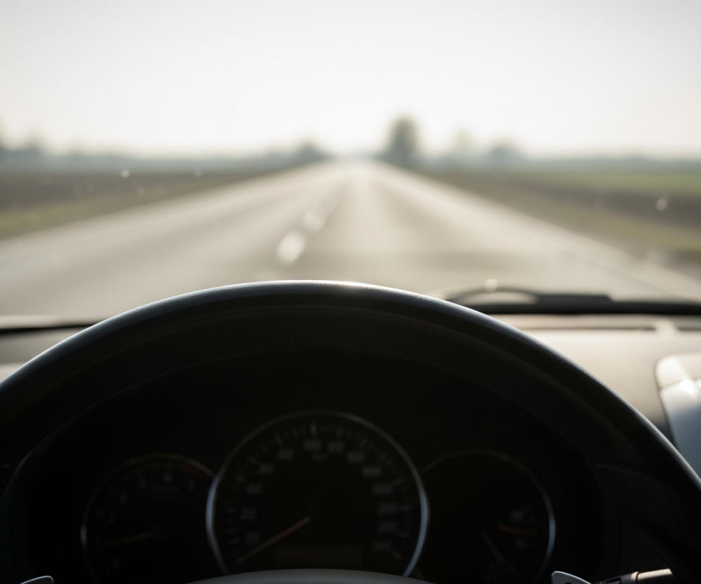 Ciemna kierownica samochodu dominuje w dolnej części kadru, zasłaniając w znacznym stopniu widok deski rozdzielczej, na której widać jedynie zarysy zegarów. Przednia szyba samochodu ukazuje rozmazany, jasny krajobraz. Długa, asfaltowa droga z białymi przerywanymi liniami pośrodku biegnie prosto w dal, znikając w jasnej, rozmytej mgiełce horyzontu. Po obu stronach drogi rozciągają się pola, a w oddali majaczą niewyraźne sylwetki drzew na tle jasnego nieba.