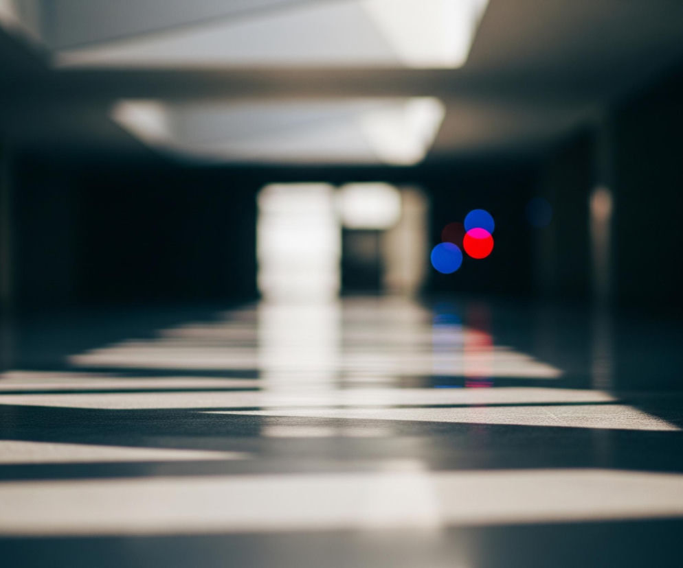 Długi, wąski korytarz z perspektywą z niskiego punktu widzenia, skupiającą się na podłodze. Na podłodze widoczne są równoległe pasy światła i cienia, rozciągające się od lewego dolnego rogu w kierunku środka, sugerując światło wpadające z góry. W tle korytarza, w dali, znajdują się rozmyte czerwone i niebieskie okręgi, przypominające światła bokeh, a także niewyraźne, jasne prostokątne kształty na suficie. Całość utrzymana jest w ciemnych, stonowanych odcieniach, z dominującymi szarościami, czerniami i ciepłymi beżami pasów światła.