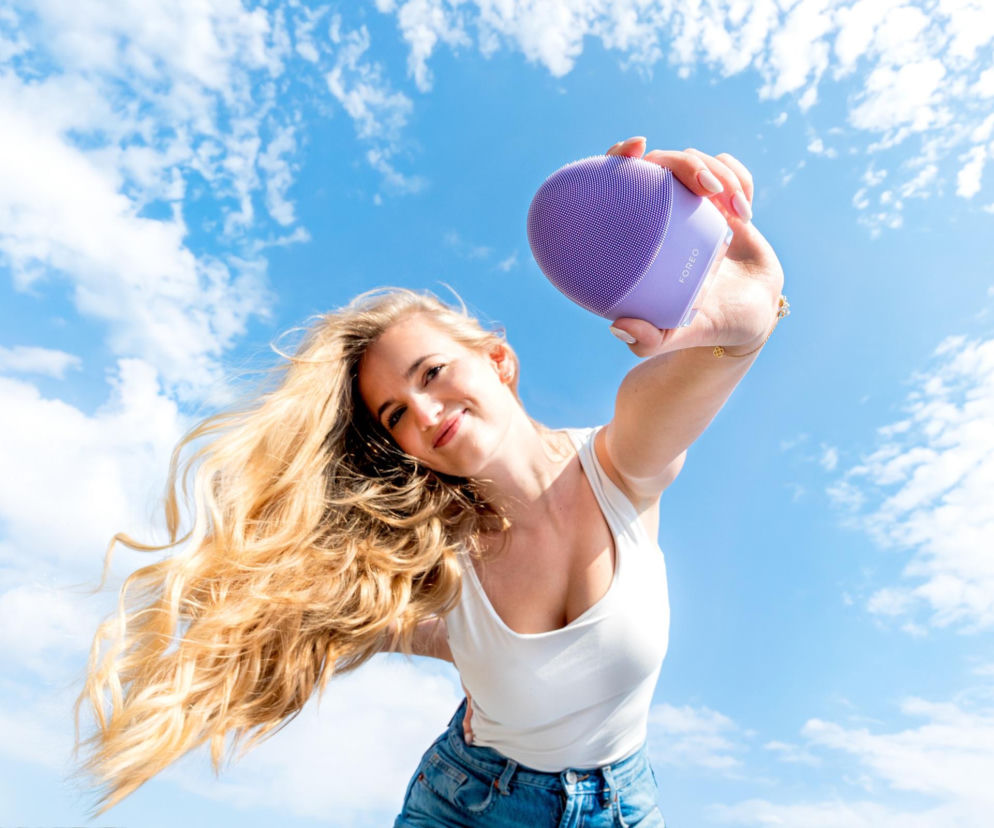 Uśmiechnięta blondynka trzyma fioletową szczoteczkę FOREO LUNA na tle nieba. O letniej pielęgnacji dowiesz się na SE.