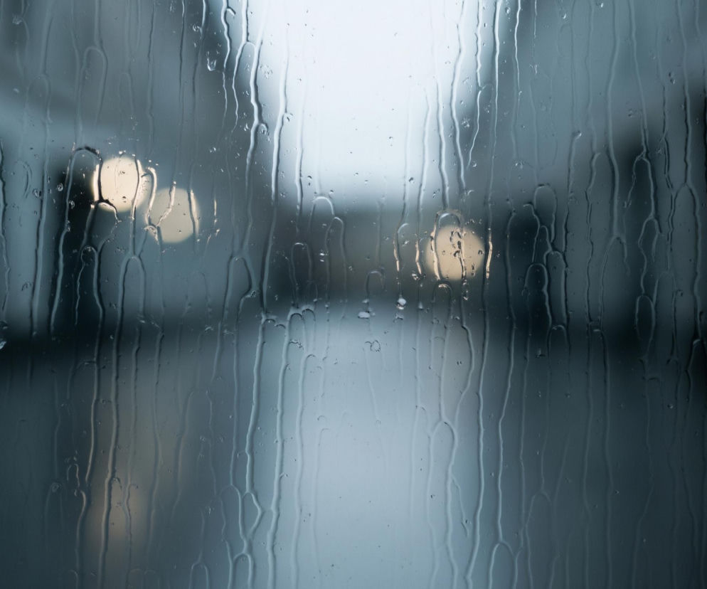 Na pierwszym planie widoczna jest mokra szyba, pokryta licznymi kroplami i smugami deszczu, które spływają pionowo, tworząc nieregularne wzory. Tło, rozmyte i nieostre, ukazuje miejski krajobraz w odcieniach szarości i błękitu, z kilkoma jasnymi, okrągłymi plamami światła w kolorze ciepłej bieli lub delikatnego żółtego, sugerującymi lampy uliczne lub reflektory pojazdów. Ogólna atmosfera obrazu jest ponura i deszczowa.
