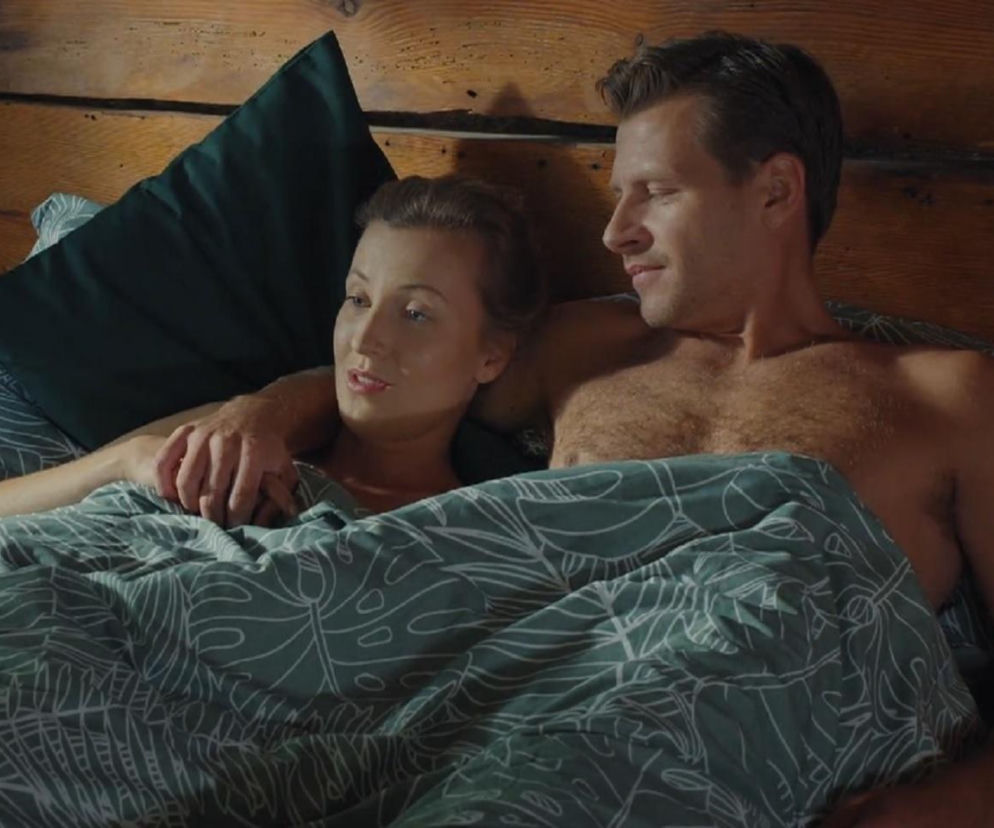 Natalka (Dominika Suchecka) i Adam Karski (Patryk Szwichtenberg) w łóżku pod zieloną pościelą, obejmujący się. Kobieta patrzy w lewo, mężczyzna w prawo, na jej twarz. Zdjęcie ilustruje scenę z serialu M jak miłość, o której możesz przeczytać więcej na Super Seriale.