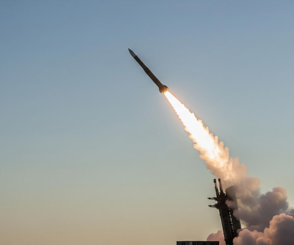 Ciemna rakieta z płonącym, jasnym ogonem dymu i ognia startuje, wznosząc się ukośnie w prawo, w stronę błękitnego nieba. Pod rakietą, w prawym dolnym rogu, widoczna jest struktura wyrzutni oraz duża ilość gęstego, ciemnego dymu, rozprzestrzeniającego się na dole po prawej stronie. Niebo, od góry w lewo, przechodzi od jasnoniebieskiego do jaśniejszego, lekko pomarańczowego odcienia u dołu.