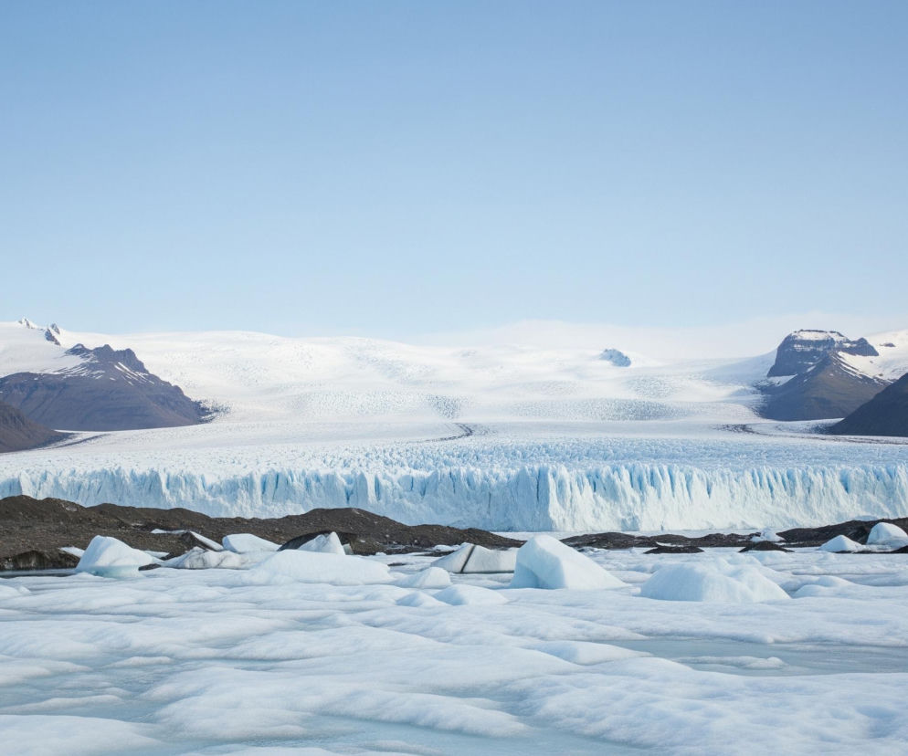 Na pierwszym planie widoczne są nieregularne bryły lodu, unoszące się na tafli zamarzniętej wody, która ma błękitno-biały odcień. Za nimi rozciąga się pas ciemnego, skalistego terenu, z kilkoma mniejszymi bryłami lodu. Dalej w tle dominuje ogromny lodowiec o wyraźnej, błękitnej fasadzie z pionowymi pęknięciami i pofalowaną, białą powierzchnią w oddali. Horyzont wyznaczają ciemne, zaśnieżone góry, a nad wszystkim rozpościera się jasne, bezchmurne, błękitne niebo.