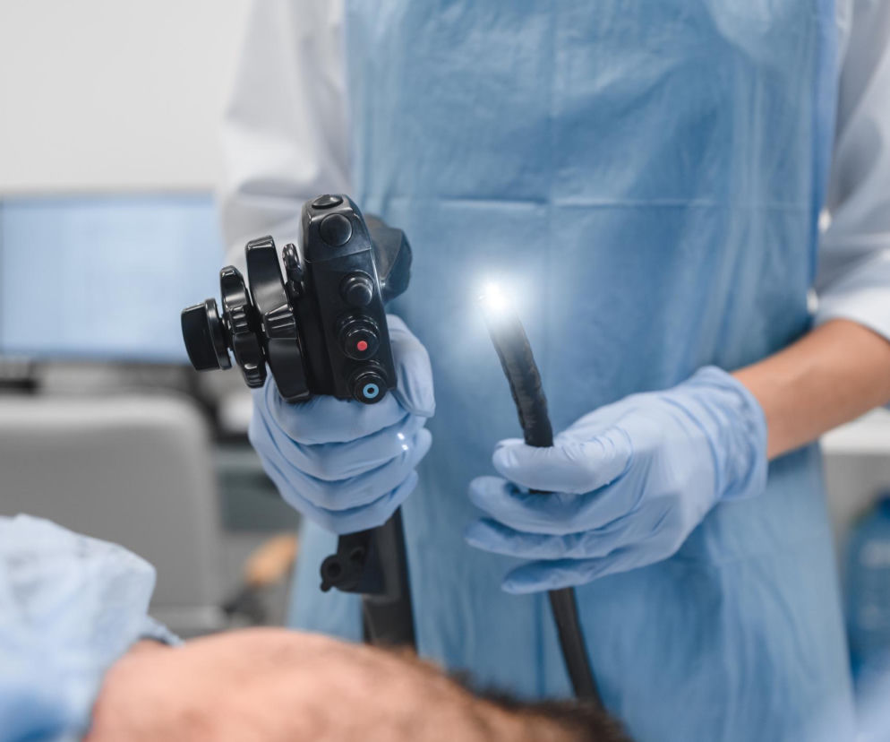 Ręce w niebieskich rękawiczkach trzymają endoskop z włączoną lampą do kolonoskopii. W tle widoczny niewyraźny monitor i zarys pacjenta. Na Poradniku Zdrowie znajdziesz informacje o tym, jak przygotować się do tego badania.