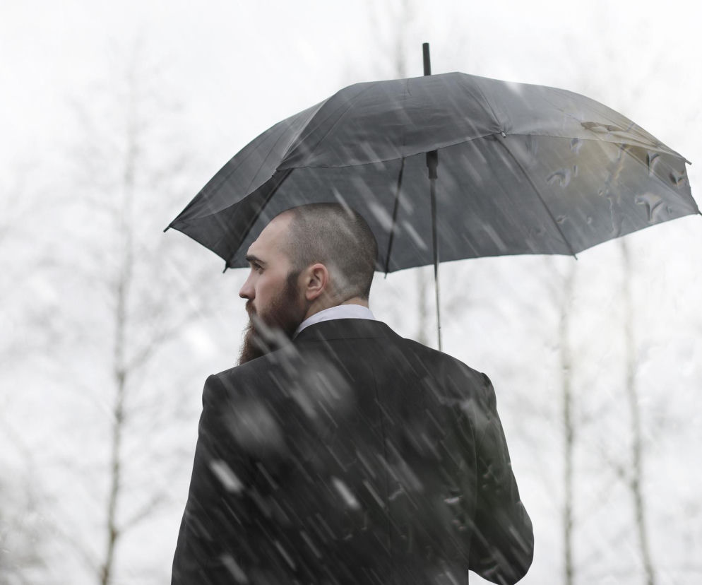 Mężczyzna z brodą w ciemnym garniturze i białej koszuli, trzymający czarny parasol podczas opadów, patrzy w lewo z zamglonym tłem, co symbolizuje wpływ nagłych zmian pogody na samopoczucie. Więcej o tym, dlaczego czujemy się fatalnie przy załamaniu pogody, przeczytasz na Poradnik Zdrowie.