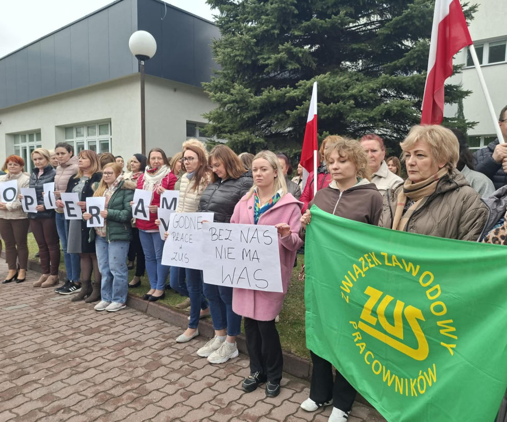 Grupa pracowników ZUS podczas protestu w Starachowicach, trzymająca transparenty z napisami Wspieram Godne Prace w ZUS i Bez Nas Nie Ma Was, oraz zieloną flagę Związek Zawodowy Pracowników ZUS. Konflikt w ZUS narasta, o czym możesz przeczytać na Super Biznes.