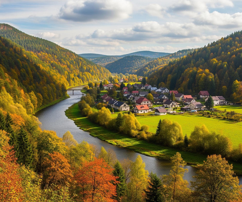 Krajobraz górski w jesiennych barwach ukazuje krętą rzekę płynącą przez dolinę otoczoną zalesionymi wzgórzami. W środkowej części obrazu, na prawym brzegu rzeki, rozciąga się wieś z licznymi domami o czerwonych i szarych dachach, otoczona soczyście zielonymi polami. Na lewym brzegu rzeki widoczny jest most łączący dwa brzegi. Niebo jest częściowo zachmurzone, z jasnymi plamami błękitu prześwitującymi między puszystymi, białymi chmurami.