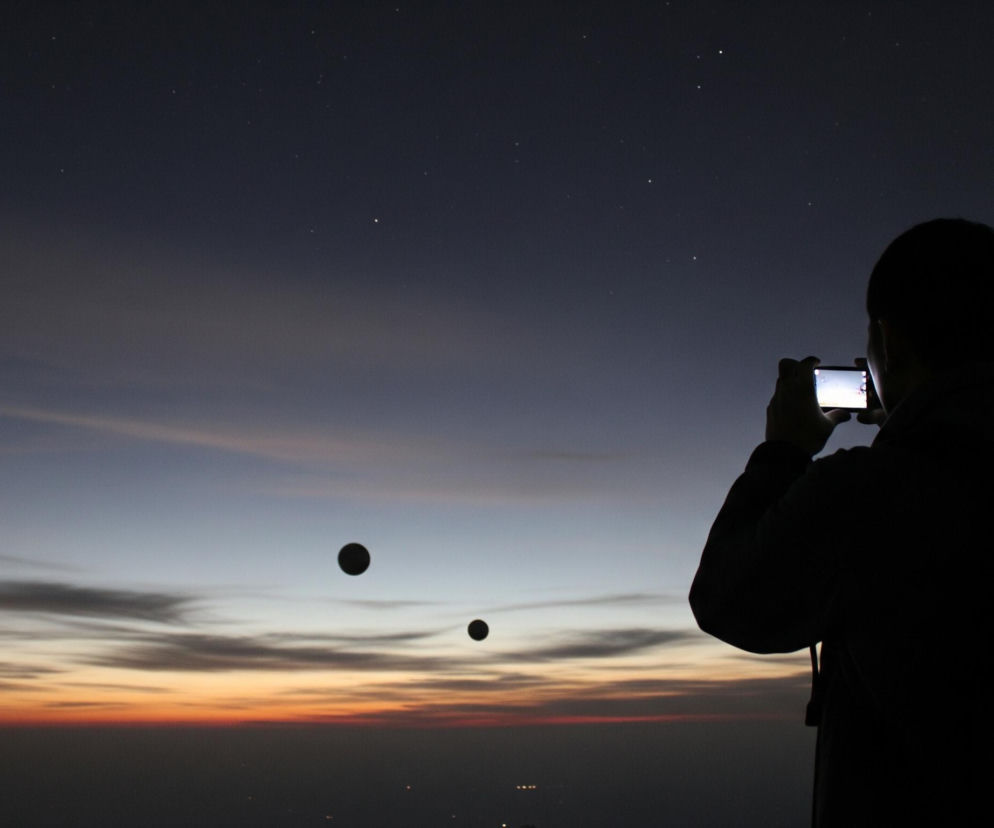 Sylwetka osoby w prawym rogu kadru, zwrócona plecami do obserwatora, trzyma w dłoniach poziomo aparat lub telefon, którego ekran świeci jasnym, białym światłem. Osoba stoi na tle nieba o przejściowych barwach, od ciemnego granatu z widocznymi gwiazdami w górnej części, przez błękit i szarość w środku, aż do pomarańczowo-czerwonej linii horyzontu. Poniżej horyzontu widać ciemny pas ziemi z rozproszonymi, drobnymi światłami. W centralnej części nieba unoszą się dwa ciemne, koliste obiekty, z których jeden jest większy i znajduje się wyżej, a drugi, mniejszy, jest położony nieco niżej i bardziej w prawo.