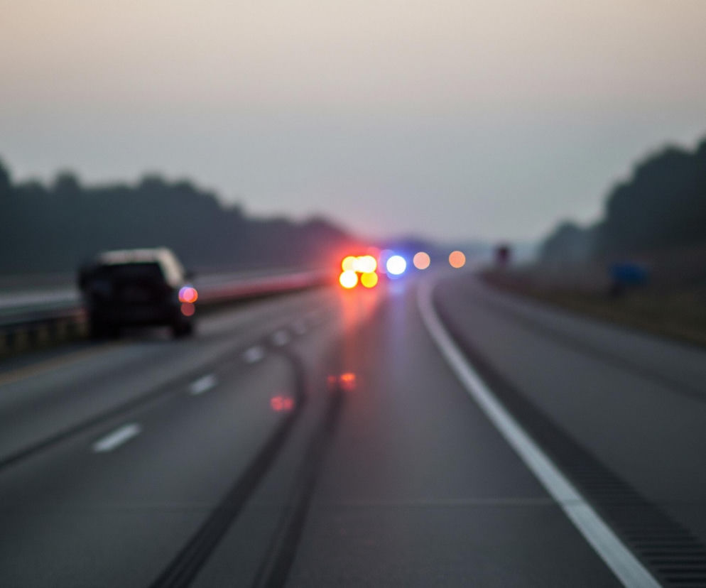 Scena przedstawia rozmyty widok z perspektywy jadącego pojazdu, ukazując autostradę o zmierzchu lub wczesnym poranku. Dwie ciemne jezdnie z białymi pasami rozdzielone są pasem zieleni pośrodku, a na nich widoczne są rozmyte światła samochodów. Ciemny samochód jedzie po lewej stronie drogi, a dalej na horyzoncie migają czerwone i niebieskie światła pojazdu uprzywilejowanego, co sugeruje sytuację awaryjną. Tło stanowią ciemne kontury drzew i jaśniejące, rozmyte niebo, co podkreśla atmosferę nastrojowego zmierzchu.