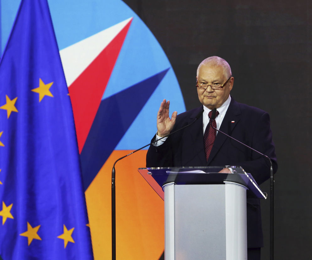 Prezes NBP Adam Glapiński podczas konferencji prasowej, z polską i unijną flagą w tle, mówi o stopach procentowych i WIBOR. Więcej na Super Biznes.