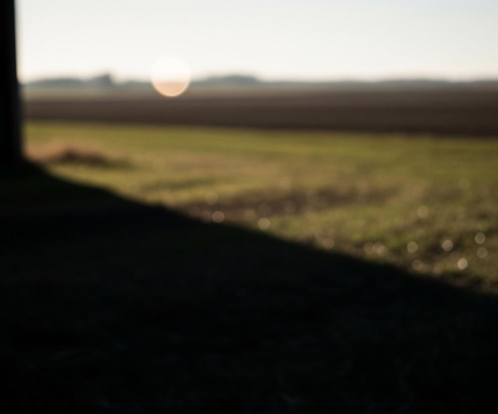Główne elementy zdjęcia to rozmyty, wiejski krajobraz oświetlony zachodzącym słońcem. Po lewej stronie widać ciemną, niewyraźną konstrukcję, przypuszczalnie fragment budynku, z jasnym okrągłym kształtem w górnej części, być może odbiciem światła. Prawa część kadru zdominowana jest przez rozległe pola, gdzie na pierwszym planie znajduje się zielone pole z rozproszonymi, jasnymi okrągłymi kształtami (bokeh), a za nim ciemniejsze, brązowe pole. Na horyzoncie widoczne są niewyraźne zarysy drzew lub wzniesień, a nad nimi jaśniejące, rozmyte słońce, dominujące w centrum góry kadru, zlewające się z jasnym, błękitno-białym niebem.