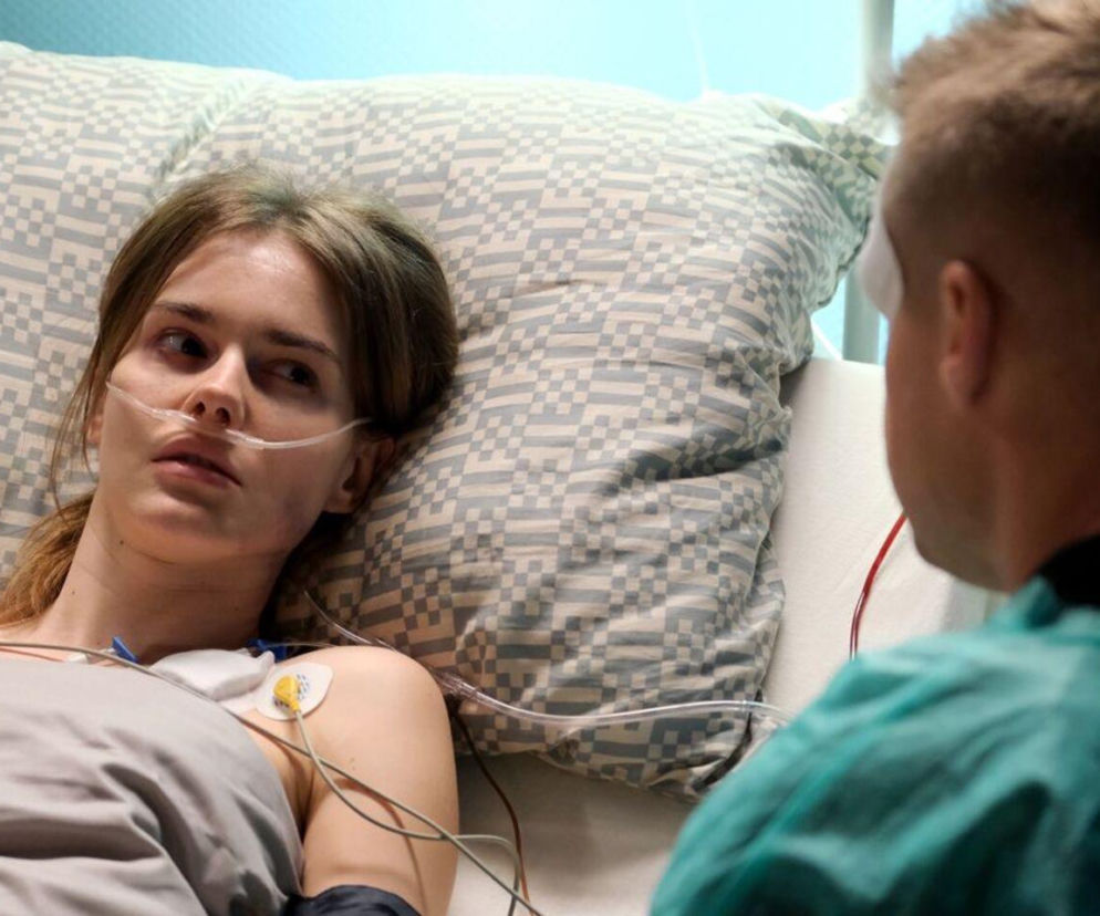 Franka Zduńska (Dominika Kachlik) leżąca w szpitalnym łóżku, podłączona do aparatury medycznej, patrzy z troską na Pawła Zduńskiego (Rafał Mroczek), który siedzi do niej tyłem, w zielonym kitlu. Scena z serialu M jak miłość dostępnego na Super Seriale.