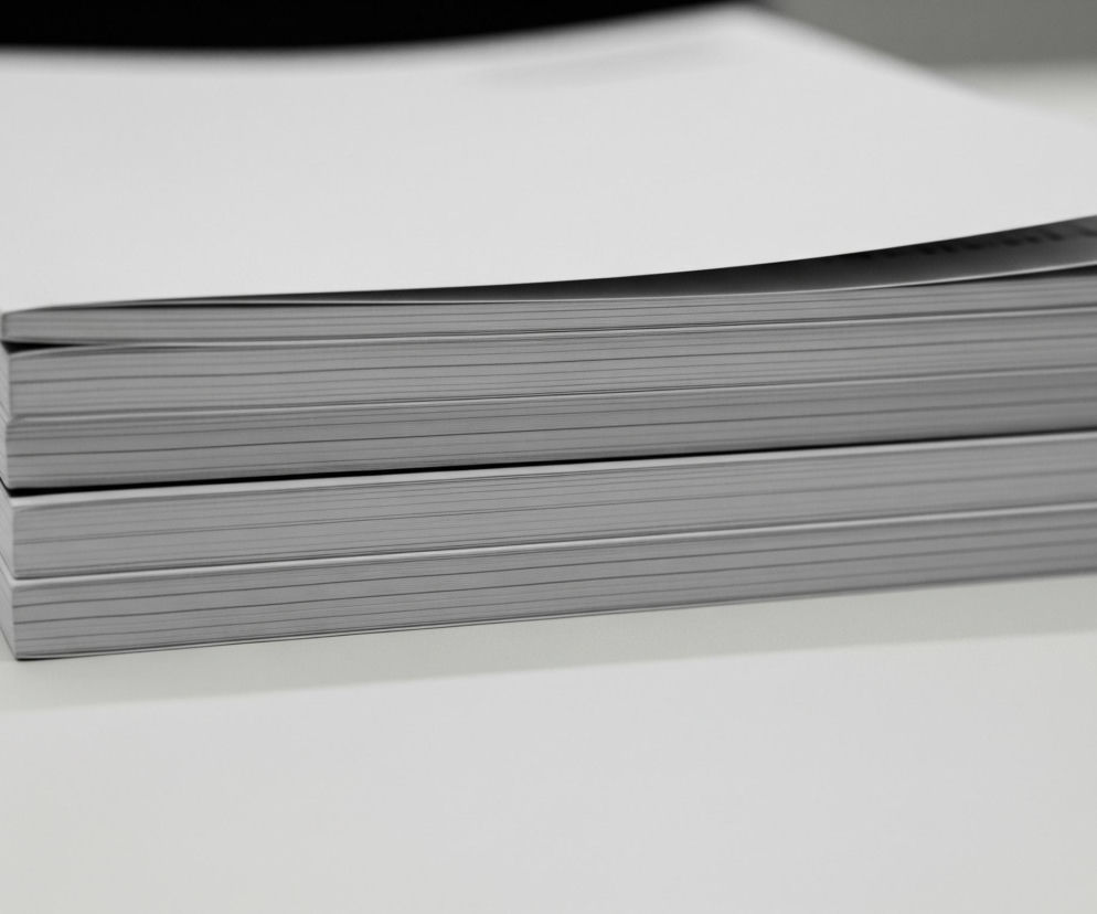 Dwa stosy białych, prostokątnych kartek papieru leżą na jasnoszarym blacie. Górny stos jest grubszy, a jego wierzch lekko się unosi, ukazując ciemny, zamazany tekst na pierwszej stronie. Tło za stosami papieru jest ciemne i niewyraźne, w odcieniach czerni i szarości.