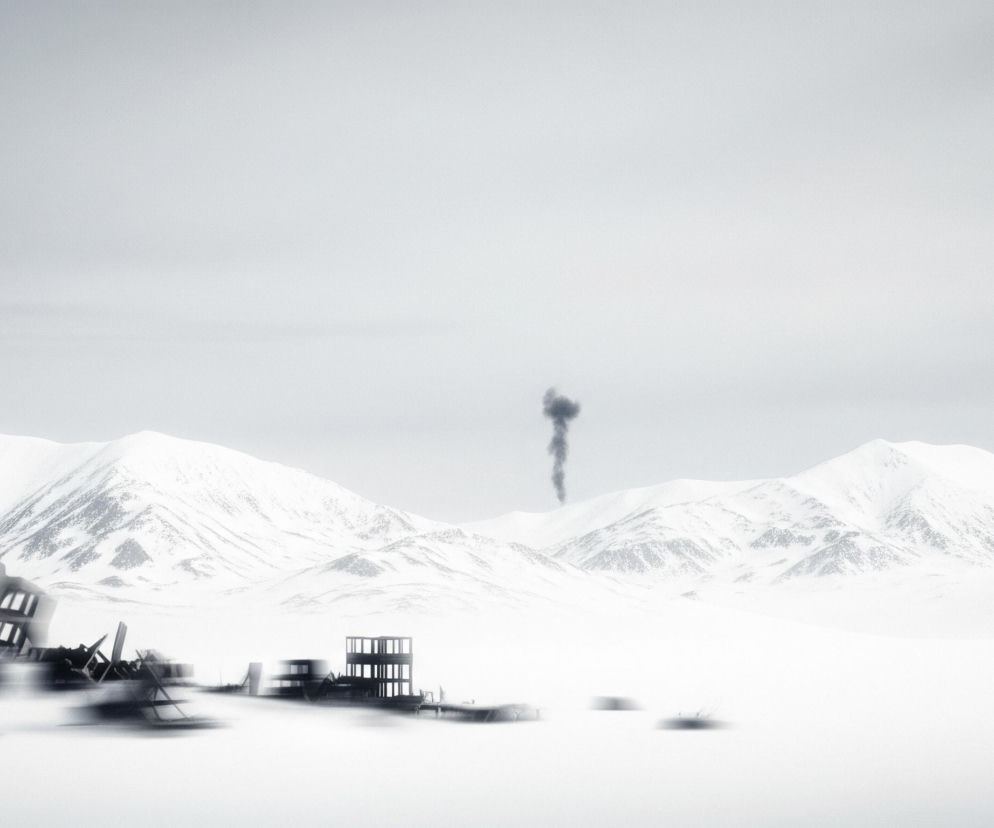 Kadruje się zimowy, górzysty krajobraz, gdzie ośnieżone szczyty wznoszą się na horyzoncie pod jasnym, zachmurzonym niebem. Na pierwszym planie, w lewej części kadru, znajdują się zrujnowane, ciemne konstrukcje budynków, ostre krawędzie i otwarte prostokątne okna kontrastują z rozmytym, białym otoczeniem. W oddali, po prawej stronie, widać rozrzucone szczątki przypominające fundamenty, również rozmyte, a pośrodku unosi się w górę ciemna smuga dymu, tworząc pionowy akcent na tle jałowego pejzażu. Cały obraz utrzymany jest w odcieniach bieli, szarości i czerni, z dominującą bielą śniegu i nieba.