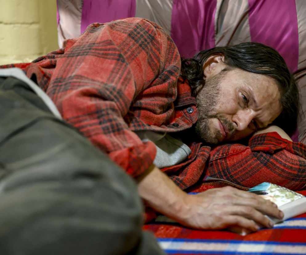 Aktor Mateusz Kościukiewicz jako Paweł w serialu Pierwsza miłość leży, opierając głowę na ramieniu, i spogląda na trzymany przedmiot. Nosi koszulę w czerwoną kratę. Scena przedstawia trudną sytuację bohatera, którego los w więzieniu w Kolumbii jest niepewny. Więcej informacji o serialu znajdziesz na Super Seriale.