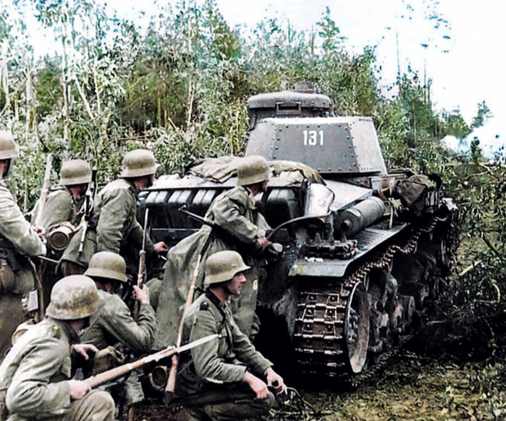 Niemieccy żołnierze w hełmach i mundurach Wehrmachtu, ukryci w zaroślach, wspierani przez czołg Panzer I z numerem 131 podczas operacji Barbarossa. W tle widać dym. Więcej o inwazji na ZSRR przeczytasz na naszym portalu.