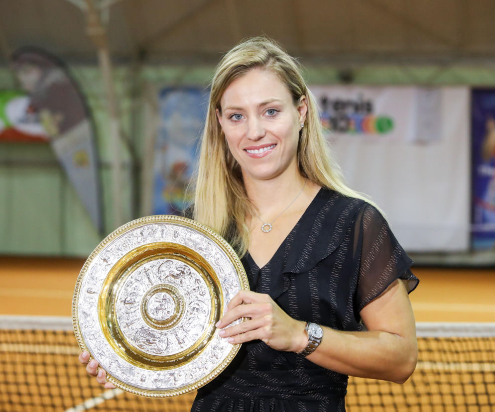 Tenisistka Angelique Kerber z blond włosami w czarnej sukience uśmiecha się, trzymając trofeum Wimbledonu. Zdjęcie, na którym widać też fragment siatki kortu, towarzyszy artykułowi o jej pożegnaniu z karierą, o czym więcej przeczytasz na naszym portalu.
