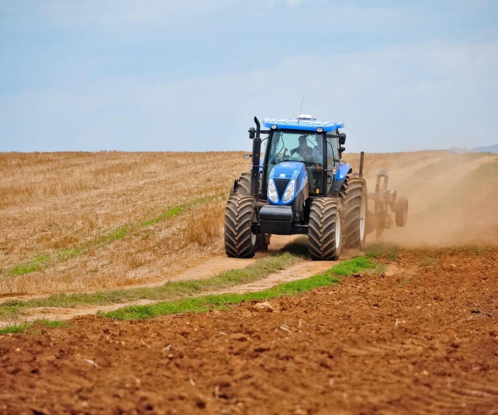 Niebieski traktor pracuje na polu, wzniecając chmurę pyłu. O terminach składania dokumentów do ARiMR przeczytasz na SE Superbiz.