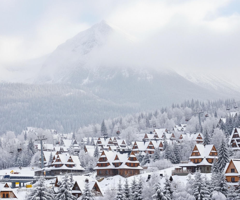 W centrum obrazu rozciąga się zimowy krajobraz z licznymi drewnianymi domami o spiczastych, ośnieżonych dachach, usytuowanymi wśród zaśnieżonych drzew iglastych. Domy są brązowe, kontrastując z bielą śniegu pokrywającego ich dachy i otoczenie. Nad zabudowaniami i lasem przebiega linia wyciągu krzesełkowego z kilkoma widocznymi wagonikami. W tle dominują masywne, ośnieżone góry, częściowo przysłonięte mgłą lub chmurami, sprawiając wrażenie majestatycznej, zamglonej scenerii.