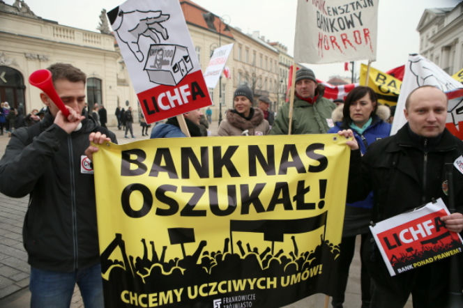 Protest frankowiczów w Warszawie, gdzie demonstranci trzymają transparent z napisem Bank nas oszukał! i transparenty potępiające lichwę. Z lewej młody mężczyzna z czerwoną tubą. Więcej o orzeczeniu TSUE korzystnym dla frankowiczów przeczytasz na Super Biznes.