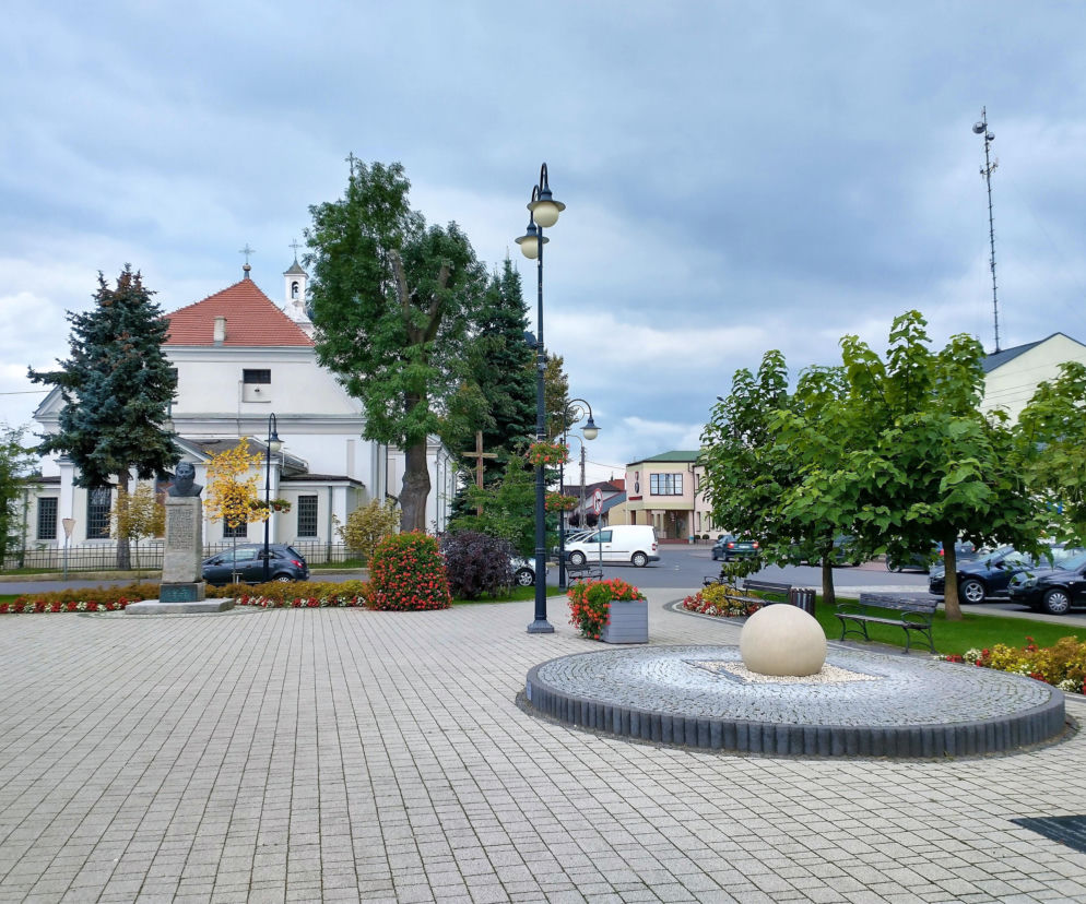 Rynek w Wolborzu z brukowanym placem, centralną kulą dekoracyjną oraz kościołem św. Mikołaja z czerwonym dachem w tle. Wokół dużo zieleni i zabytkowe kamieniczki, świadczące o bogatej historii tego jednego z najstarszych miast w woj. łódzkim, o którym przeczytasz więcej na naszym portalu.