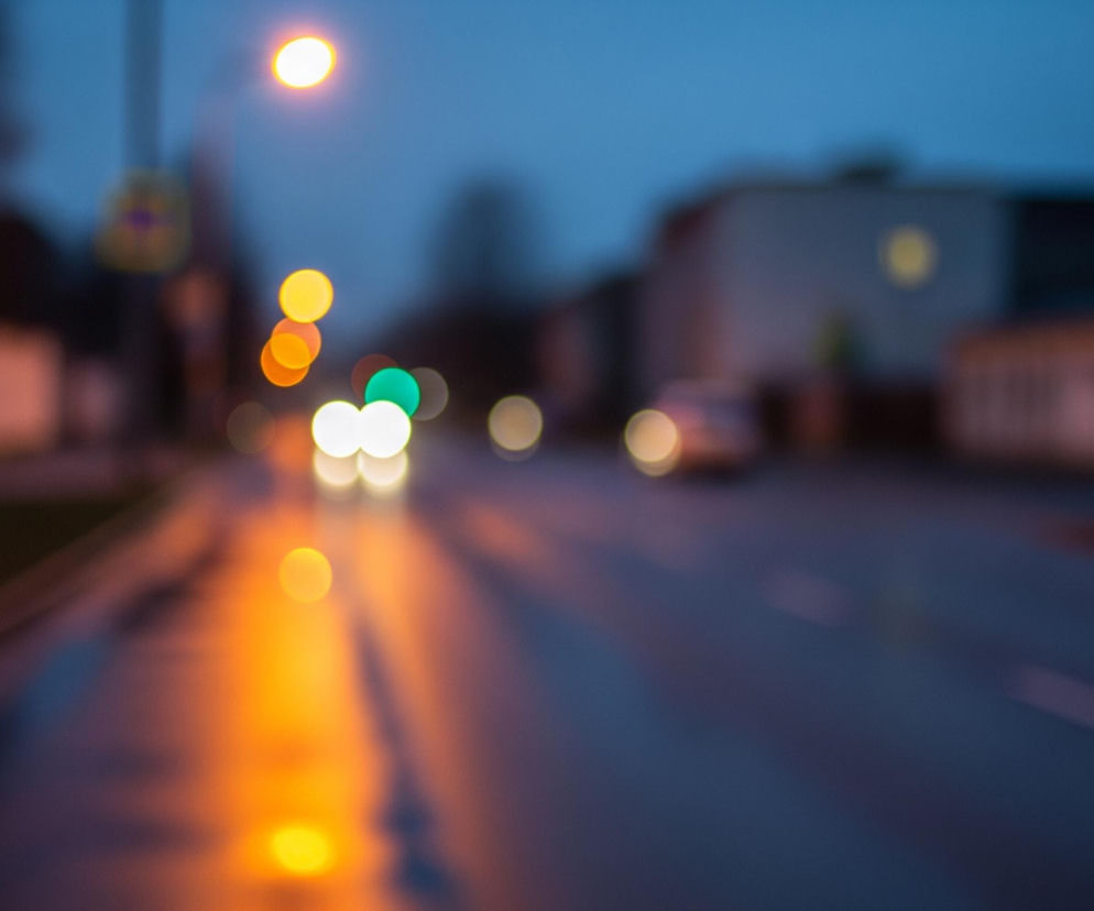 Miejski krajobraz nocą lub o zmierzchu, utrzymany w stylistyce bokeh, z rozmytymi światłami w tle i na pierwszym planie. Na pierwszym planie widoczna jest mokra nawierzchnia drogi, odbijająca pomarańczowe i białe światła pojazdów oraz latarni ulicznych, tworząc długie, rozmyte smugi. W centrum kadru znajdują się rozmyte światła samochodów, w tym zielone światło przypominające sygnalizację świetlną, a także szereg pomarańczowych i białych okręgów. W tle widać zarysy budynków i drzew pod ciemnym, niebieskawym niebem, a także pojedyncze, jasne punkty świetlne.