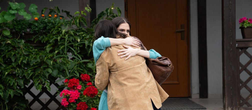 Basia i Agnes z serialu M jak miłość przytulają się na pożegnanie przed domem, stojąc na schodach. Basia w turkusowej bluzie, Agnes w beżowej kurtce. Scena ukazuje smutek i bliskość sióstr, które żegnają się w pośpiechu. Więcej o pożegnaniu bohaterek na Super Seriale.