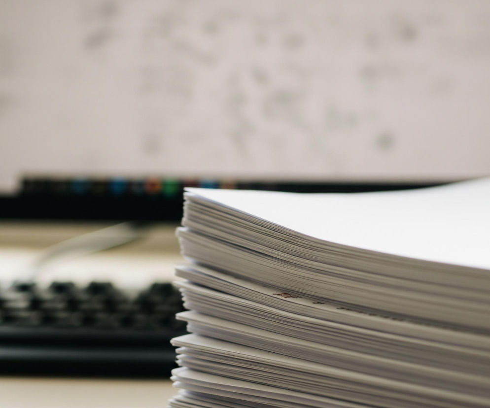 Na pierwszym planie, z prawej strony, znajduje się wysoki stos białych kartek papieru, ułożonych jedna na drugiej, tworząc nieregularny, lekko zakrzywiony kształt. Kartki są gładkie, a na niektórych z nich widoczne są drobne, niewyraźne brązowe lub czerwonawe znaki. Tło, rozmyte i nieostre, ukazuje fragment monitora komputera z widocznym na ekranie jasnym, niezdefiniowanym tekstem lub grafiką, oraz część czarnej klawiatury z rzędami klawiszy.