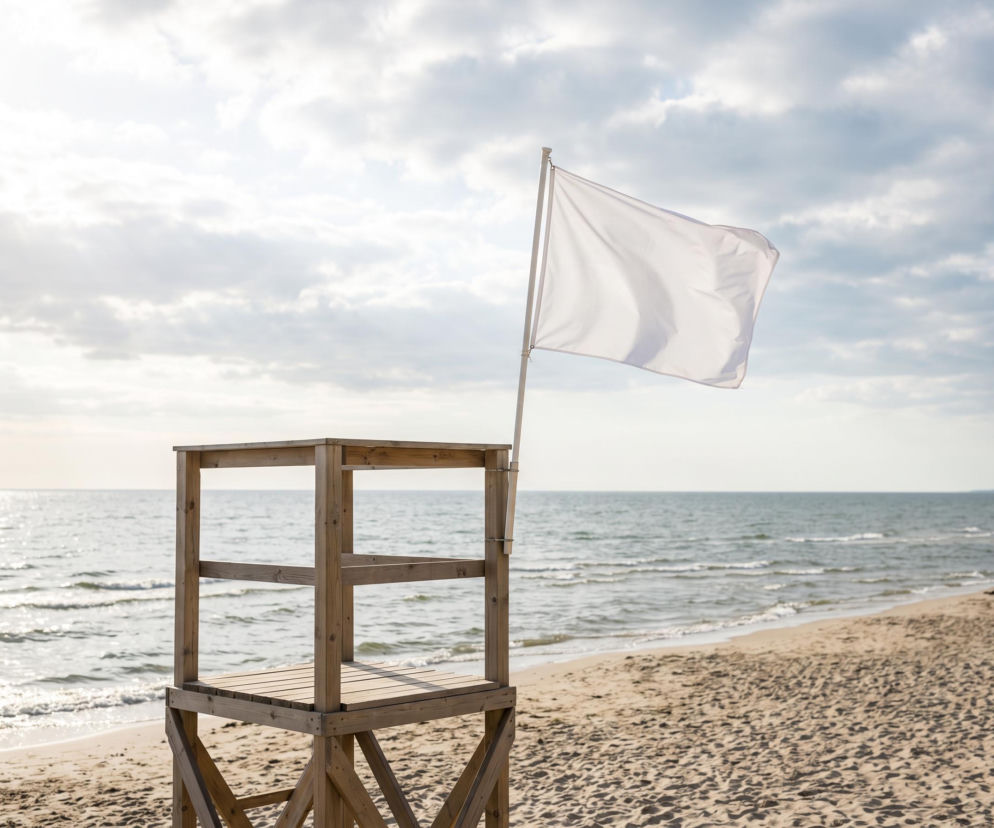 Biała flaga na wieży ratowniczej przy pustej plaży. O bezpieczeństwie kąpieli w Chłopach informuje SE Szczecin.